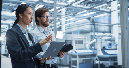 Two Multiethnic Engineers Standing in Office, Using Laptop and Tablet Computers at an Electronics...