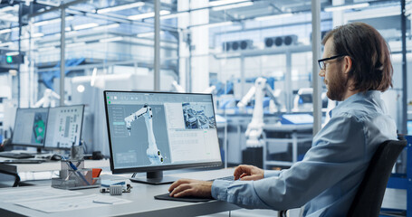 Portrait of a Young Factory Manager Working on Desktop Computer in Industrial Manufacture with Autonomous Assembly Robots. Production Line Making Heavy Machinery Parts in the Background