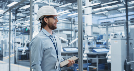 Young Electronics Manufacture Engineer Smiling, Looking at Production Line. Portrait of a Man Wearing a Protective White Hard Hat. Specialist Monitoring Artificial Intelligence Systems at a Factory
