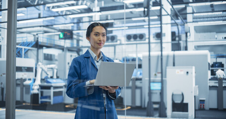 Fototapeta premium Electronics Manufacturing Technician in a Blue Coat Working on Laptop Computer at an Assembly Plant. Asian Female Specialist Developing, Maintaining, Setting Up Software for Automated Production