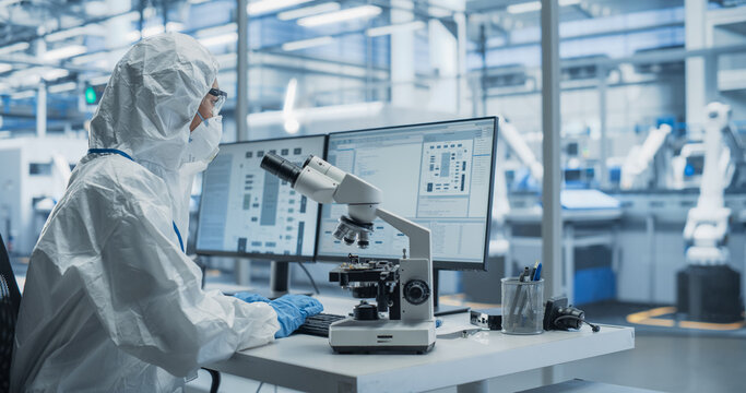 Research Factory Cleanroom: Engineer Wearing Coveralls, Gloves and Goggles while Using Microscope to Inspect a Microchip. Scientist Developing Advanced Electronics Component for the Precision Industry