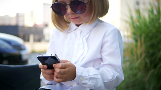 A little girl sitting on a bench and smiling, intently using her cell phone. A young girl writes a message in a social network on her phone while perched on a park bench. 
