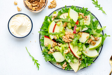Delicious crispy vegan salad with green and red apples, fresh celery, lettuce, arugula and walnuts on plate, white background, top view