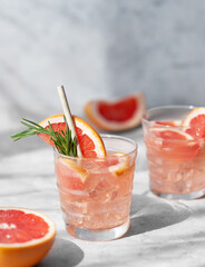 Grapefruit mojito cocktail with rosemary and ice in a two glasses on a light background with fresh citrus fruits and shadow.