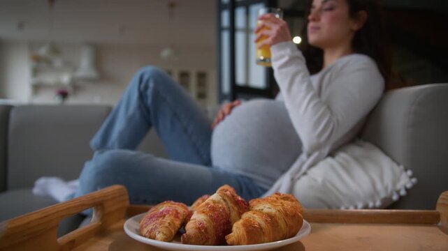 Caucasian girl European pregnant woman mom female lady enjoy pregnancy big tummy at home couch sofa relaxing drinking healthy fresh orange tropical juice eating croissant bun nutrition food delivery