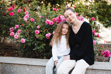 Fototapeta premium Young smiling woman holding child girl 5-6 year old over blooming rose flowers at city street outdoor. Childhood.