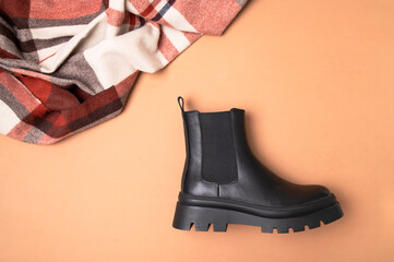Black leather classic women's boots and checkered autumn wool scarf on beige background top view flat lay. Minimalistic layout, autumn footwear advertisement. Fashionable stylish boots