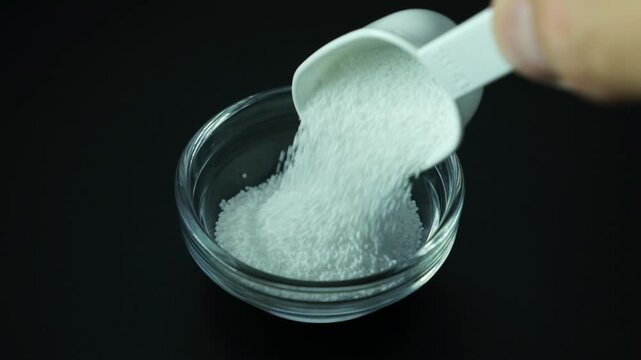 Granulated Sodium percarbonate powder or sodium carbonate peroxide is poured from a measuring spoon into a glass bowl. Video, Slow motion, close-up. 