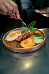 close up in a professional kitchen on a clean metal table woman chef collects English breakfast on a round wooden plate