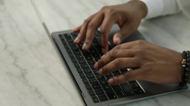 Cropped hands of black person typing on keyboard of portable computer placed on light marble surface. Busy freelancer writing email with terms of cooperation for client