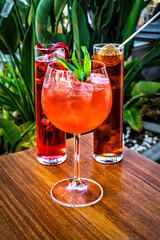 Close-up of glass filled with Aperol Spritz. At the background two red cocktails served in highball glasses filled with a lot of ice.