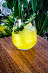Stemless tulip glass filled with ice and a vibrant yellow cocktail, garnished with a fresh cucumber slice and a sprig of rosemary. Fresh, herbal touch