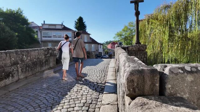 Caldas de Reis, Pontevedra - Galicia, Spain. Pilgrims walking on a historic stone bridge adorned with a cross, part of the Camino de Santiago route as it passes through the town.