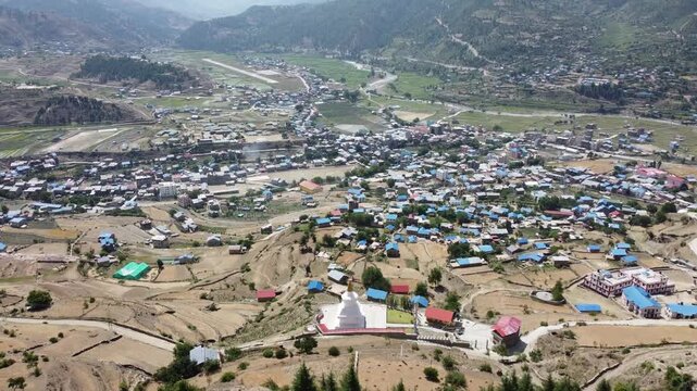Aerial view of Jumla Bazar, a town in western Nepal surrounded by mountains. The town's buildings are spread across terraced fields, with a small river running through the valley.