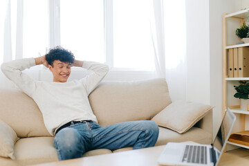 Fototapeta premium Young man relaxing on a sofa in a bright room, smiling and enjoying some leisure time in a cozy home setting A laptop rests on the table nearby, showcasing a casual atmosphere