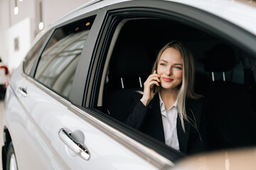 Happy attractive blonde young woman customer sitting inside new luxurious car, talking mobile phone, happy buying auto in dealership. Concept of choosing and buying new auto at showroom.