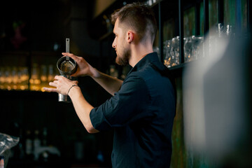 A young bartender in a black shirt prepares an alcoholic drink by pouring alcohol from  shaker in a...