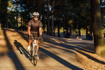 Fototapeta premium A cyclist in a helmet and sunglasses rides along a sun-dappled forest path, casting a long shadow.
