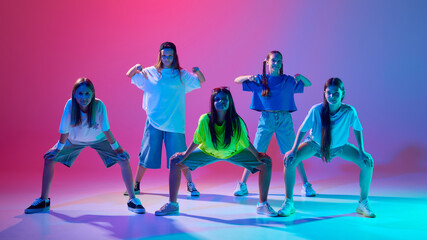 Stylish children, young girls standing and posing in dynamic pose, hip hop dancers against gradient pink purple background in neon. Concept of modern dance styles, youth culture, active lifestyle