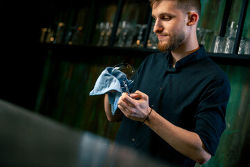 in a night bar a young bartender with a beard in a black shirt rubs a martini glass with a blue...