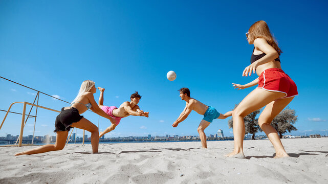Players leap into the air, battling it out during a beach volleyball game. Group of friends having fun on beach by the river. Concept of sport, summer activity, leisure games, friendship