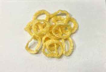 The image shows a group of light yellow, ring-shaped snacks arranged on a white surface, suggesting a crunchy texture and savory flavor.