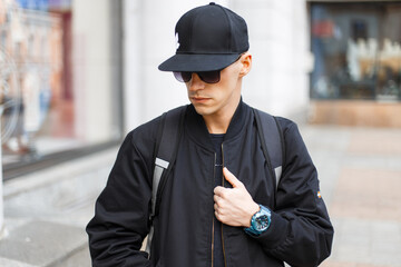 Handsome man with black cap in fashionable black jacket with backpack walking in city. Stylish guy in autumn clothes