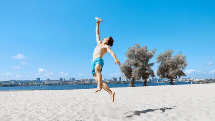 Man in beach shorts playing frisbee on beach by the river under clear blue sky. Dynamic game,...