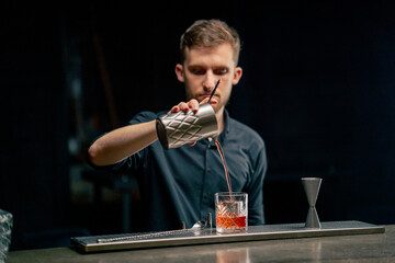 in a bar on a black background a handsome young bartender in a black shirt pours an alcoholic drink...