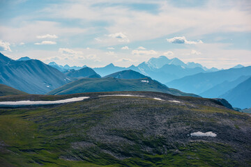 Alpine aerial top view to sunlit big green hill and large rocky mountains silhouettes in bright sun under clouds in blue sky. Scenic layered landscape with high snowy mountain far away in sunny day.