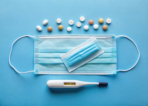 A arrangement of essential healthcare items, including capsule tablets, a face mask, and a digital thermometer, posed on a calming blue background.