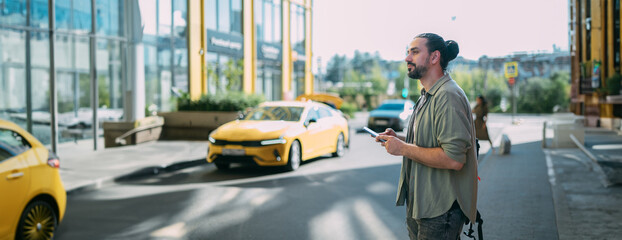 A young Caucasian man with a phone calls a taxi through an app on a city street. The guy uses an application on his smartphone to call a taxi