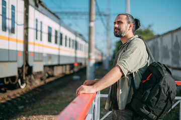 A man is waiting for a train on the platform. A young guy, a passenger with a backpack is standing on the platform waiting for the train