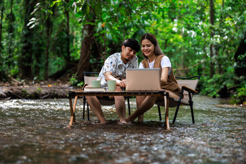 couple enjoying sitting on wooden chairs using laptop computer and drinking coffee together in middle of a stream in tropical forest on vacation, freelance and relaxing concept..