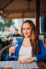 Young beautiful adult woman wearing jacket sitting next to the table in street cafeteria and hold glass of sparkling wine drink romantic date or businesswoman rest