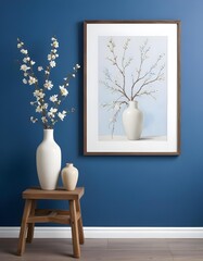 A white vase with branches and flowers on a wooden stool in front of a blue wall with a white framed artwork.