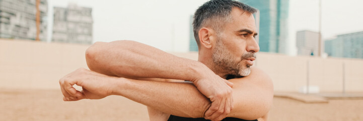 Obraz premium Athletic mature man stretches arm muscles while standing on the beach in the morning
