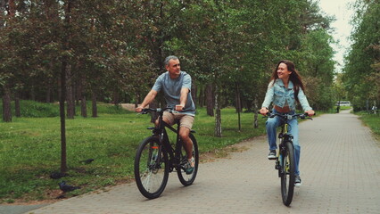 Couple enjoys biking together in park, smiling and talking while riding on tree-lined path. Concept of outdoor activities, bonding, and healthy lifestyle
