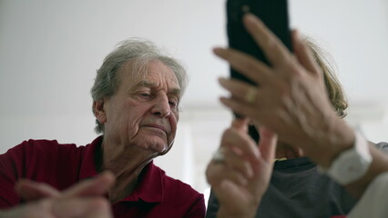 Elderly couple using smartphone together, man looking at screen, woman explaining, engaged in technology, senior lifestyle, digital learning, modern devices, bonding over gadgets, indoor scene