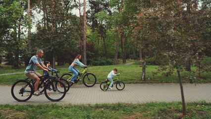 Family enjoying bike ride in park, with parents and children cycling together on paved path surrounded by trees. Concept of family bonding, outdoor activities, and healthy lifestyle