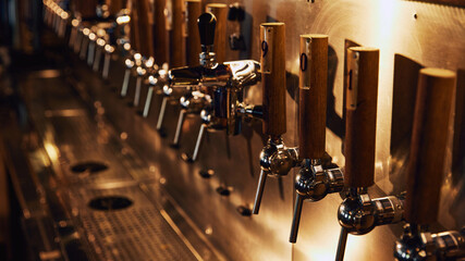 Row of shiny beer taps with numbered wooden handles, mounted on metallic wall with metallic drip tray in front. Concept of beer, brewery, pub, Oktoberfest, traditions