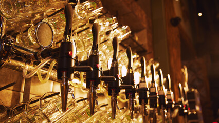 Row of shiny beer golden taps with handles, set against wall with rows of empty glasses hanging from it. Pub aesthetics. Concept of beer, brewery, pub, Oktoberfest, traditions