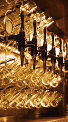 Close-up of multiple shiny beer taps with handles, set against blurred background with rows of empty glasses hanging from it. Luxury restaurant aesthetics. Concept of beer, brewery, pub, traditions