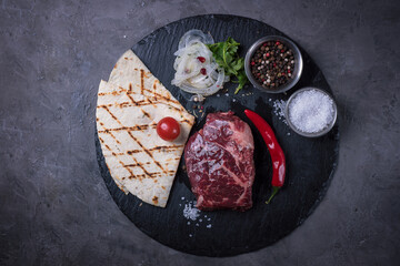 Raw chuck roll steak with pita bread, onion and spices on black stone board on grey background, top view