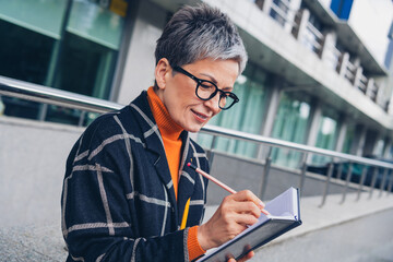 Portrait of nice aged woman pencil write take notes notepad walk downtown street outdoors