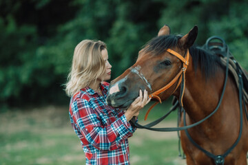 Fototapeta premium Happy blonde with horse in forest. Woman and a horse walking through the field during the day. Dressed in a plaid shirt and black leggings.
