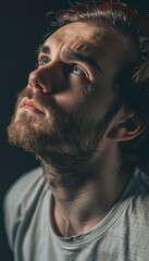 Fototapeta premium Moody portrait of a pensive young man with a beard looking up into the light, captured with dramatic shadows and a contemplative expression.