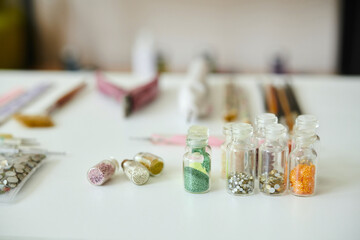 Close-up view of small transparent bottles filled with colorful beads arranged neatly on white table, with various art supplies blurred in background creating an artsy atmosphere