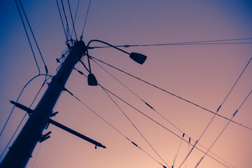 A street lamp, standing against the backdrop of the sunset sky, in the twilight of summer. Long wires extend from it, connecting it to the city's infrastructure.