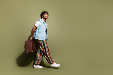 Full size photo of handsome young guy walk hold retro valise enjoy travel wear trendy jeans outfit isolated on khaki color background